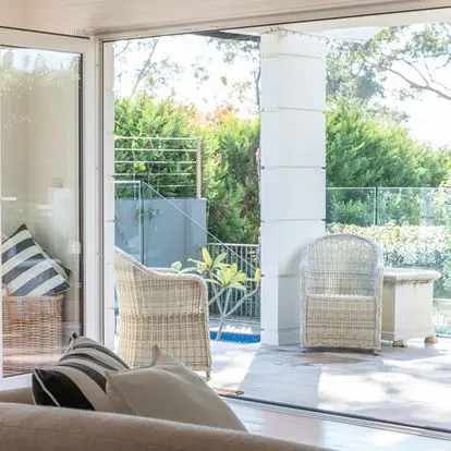 A view from inside a room showing a patio with two wicker chairs and lush greenery outside.